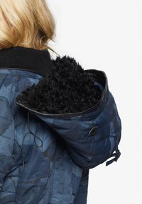 En blond person iført en blå mønstret quiltet jakke med en stor sort faux fur foret hætte, set fra bagenden.