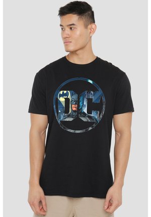 Mann trägt schwarzes T-Shirt mit kreisförmigem DC-Logo, das Batman und eine Stadtsilhouette mit dem Bat-Signal im Hintergrund zeigt.