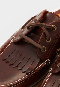 Zapatos de barco de cuero marrón con detalle de flecos, ojetes de latón y cordones marrones. Presenta costuras blancas a lo largo de los bordes para un contraste.