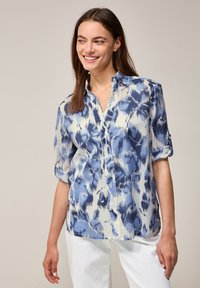 Blusa de patrón azul y blanco con cuello y mangas enrolladas, hecha de una tela ligera con una textura suave.