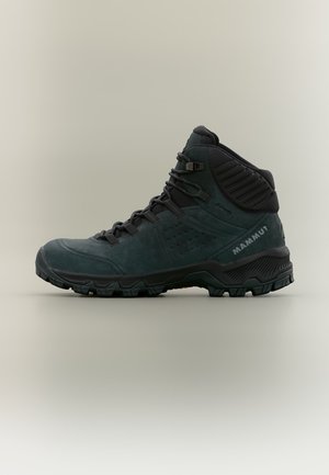 Scarponcino da trekking alto verde scuro con suola nera, lacci robusti, supporto imbottito per la caviglia e nome del marchio "Mammut" sul lato del tallone.