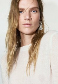 Suéter de punto rosa claro con textura fina, combinado con pendientes geométricos transparentes. La modelo tiene el cabello rubio largo y ondulado y maquillaje neutro.