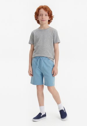 Rudovlasý chlapec stojí, oblečený v sivom tričku, svetlomodrých šortkách so sťahovacie šnúrkou, bielych ponožkách a tmavomodrých slip-on topánkach na bielom pozadí.
