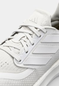Hvit idrettssko med en teksturert overdel i stoff, flate lisser og tre hvite stripe-detaljer. Rund tå og polstret krage.