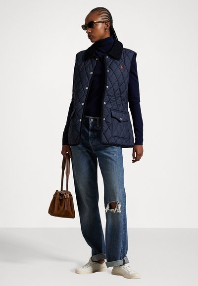 Marine blauwe gewatteerde vest met zwarte kraag, voorkeurszakken, gecombineerd met donkergroene versleten jeans en witte sneakers, terwijl ze een bruine handtas vasthoudt.