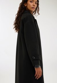 Długie czarne płaszcza wykonane z grubej tkaniny, z klapami, bocznymi kieszeniami i mankietami zapinanymi na guziki. Gładka struktura z dopasowanym krojem.