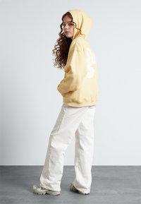 Sudadera amarilla con un bolsillo frontal y un gráfico blanco en la parte posterior, combinada con pantalones blancos holgados y zapatillas grises.