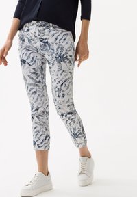 Pantalon blanc avec un motif de feuilles bleues, présentant une coupe slim et une longueur raccourcie. Le matériau semble lisse avec un léger brillant. Associé à des baskets blanches.