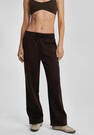 Kobieta ubrana w ciemnobrązowe szerokie spodnie dresowe z troczkiem i beżowe sneakersy, tułów widoczny od połowy klatki piersiowej do stóp na jednolitym tle.