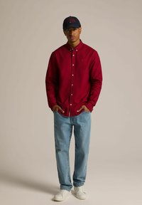 Czerwona koszula z długim rękawem zapinana na guziki, jasnoniebieskie dżinsy i białe trampki. Koszula ma kołnierzyk oraz detal z logo. Czapka jest granatowa.