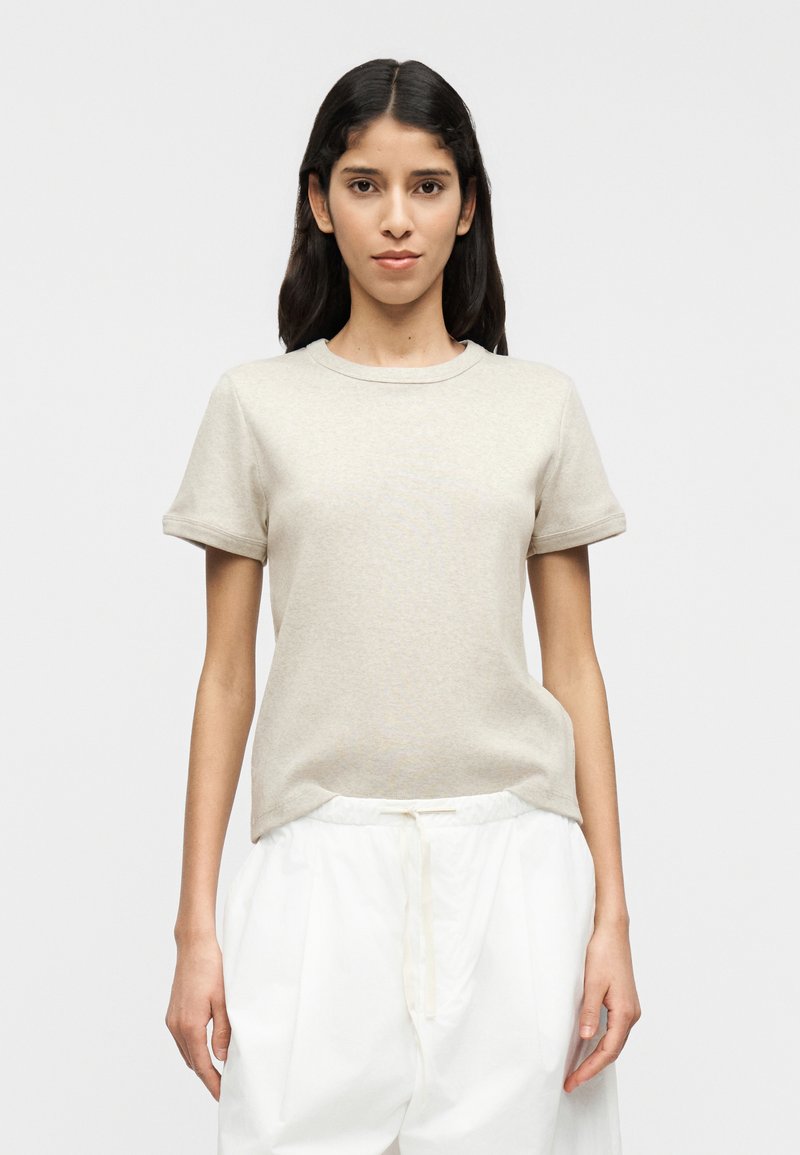 Μπεζ κοντομάνικο πουκάμισο από μαλακό, υφασμάτινο υλικό με στρογγυλή λαιμόκοψη, συνδυασμένο με άνετο λευκό παντελόνι, παρουσιάζεται σε ουδέτερο φόντο.