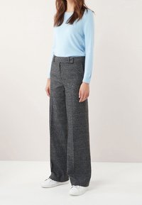 Femme portant un haut à manches longues bleu clair, un pantalon large à carreaux gris taille haute et des baskets blanches, se tenant devant un fond uni.