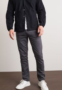 Giacca leggera nera, pantaloni grigi slim-fit e sneakers bianche. La giacca presenta una chiusura con bottoni e polsini elasticizzati; i pantaloni hanno una texture liscia.