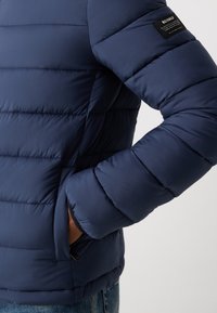 Veste d'hiver bleue marine avec des coutures horizontales étroites, poche latérale zippée et étiquette en tissu sur la manche gauche. Texture lisse et légère.