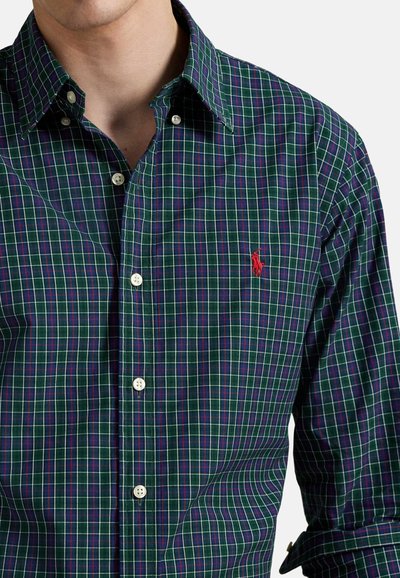 Camisa de hombre con botones, presenta un patrón de cuadros verde con líneas moradas y blancas, un pequeño logo rojo y botones blancos.