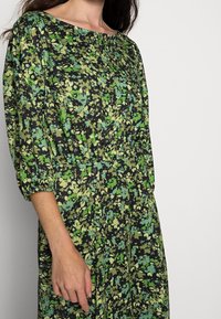 Kukkamekko mustassa ja vihreässä, jossa on löysä istuvuus, frillat hihat ja pyöreä pääntie teksturoidulla pinnalla.