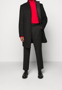 Czarny płaszcz z wełny z wysokim kołnierzem, z sześcioma guzikami z przodu, w zestawieniu z dopasowanym czerwonym golfem i czarnymi spodniami. Czarne buty wizytowe.