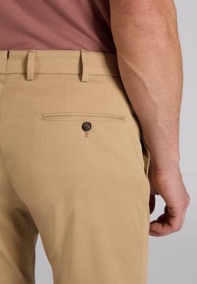 Pantalon beige d'homme avec poche arrière passepoilée fermée par un bouton foncé et passants de ceinture visibles, main détendue sur le côté.