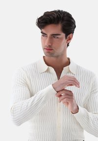 Cremfarbendes Langarmshirt mit vertikaler Rippenstruktur und dezenten Linienmustern. Verfügt über einen klassischen Kragen und eine Knopfleiste.