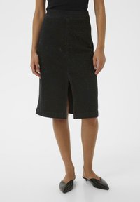 Žena v čiernej sukni po kolená s jemným leskom a predným rozparkom, v kombinácii s čiernymi špicatými slip-on obuvami.