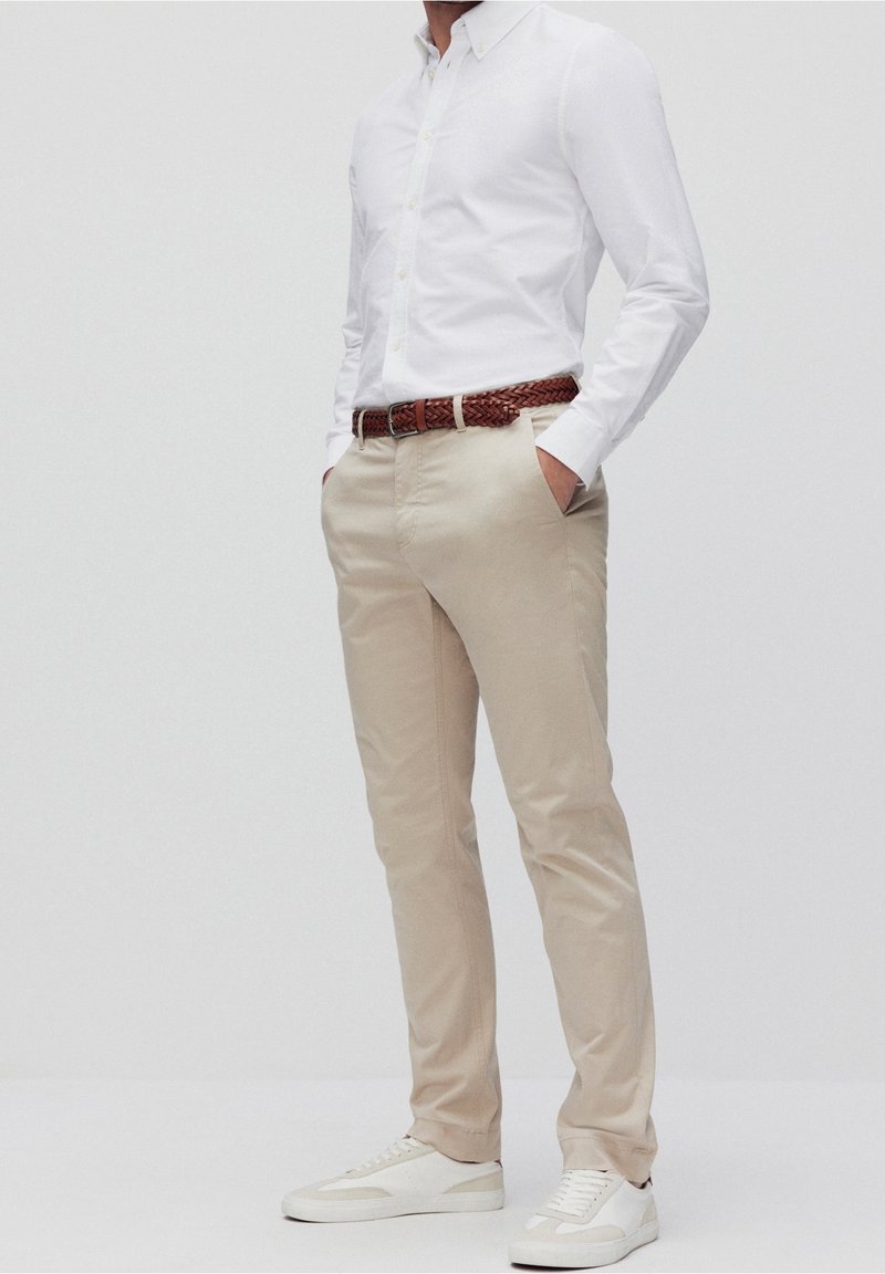 Personne portant un pantalon beige, une chemise blanche boutonnée, une ceinture tressée marron et des baskets blanches, debout les mains dans les poches.