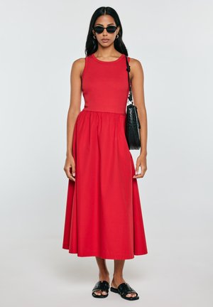 Kobieta w czerwonej sukience midi bez rękawów, czarnych okularach przeciwsłonecznych, czarnej plecionej torbie na ramię i czarnych sandałach wsuwanych stoi na tle jednolitego tła.