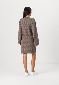Robe tricotée dans des tons marron et bleu, mi-longue, avec des manches longues et une coupe décontractée. Portée avec des baskets blanches, texture minimale visible.