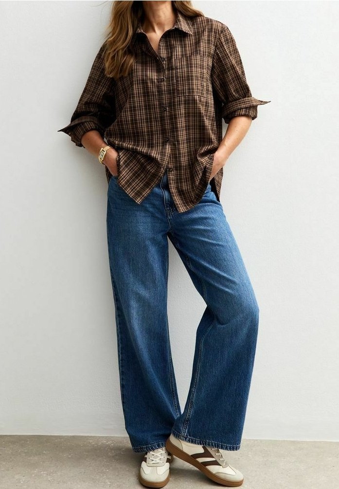 Femme portant une chemise à carreaux marron boutonnée, un jean bleu ample et des baskets beiges avec des rayures marron, debout les mains dans les poches.