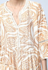 Robe à motifs beige et blanche avec un décolleté en V, des détails de boutons et des manches trois quarts. Fabriquée en tissu léger.