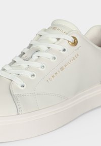 Άσπρο χαμηλό sneaker Tommy Hilfiger με άσπρα κορδόνια, λεπτομέρειες με χρυσά νούμερα και λογότυπο της μάρκας με χρυσά γράμματα στην πλαϊνή επιφάνεια.