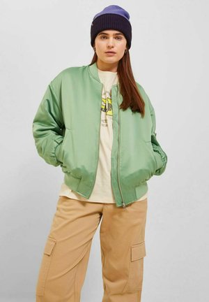 Giacca bomber in satin verde chiaro con polsini a costine, zip frontale e tasche laterali, indossata sopra una maglietta grafica color crema e pantaloni cargo beige.