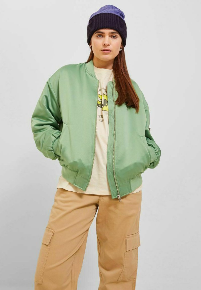 Giacca bomber in satin verde chiaro con polsini a costine, zip frontale e tasche laterali, indossata sopra una maglietta grafica color crema e pantaloni cargo beige.