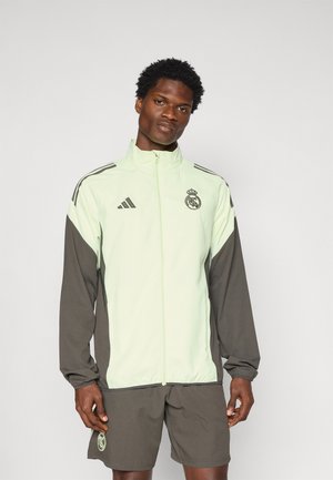 Veste Adidas vert clair avec des manches gris foncé, fermeture éclair et logo du Real Madrid sur la poitrine. Texture lisse et coupe athlétique.
