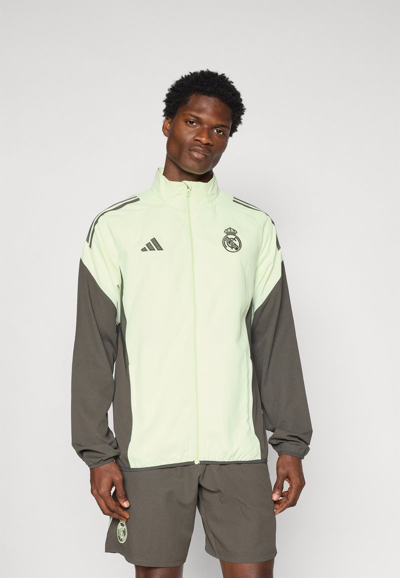 Svijetlozeleni Adidas jakna s tamnosivim rukavima, zatvaranje na zatvarač i logo Real Madrida na prsima. Glatka tekstura i atletski kroj.