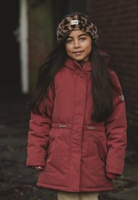 Rode gewatteerde jas met verstelbare taille, zijzakken en een bruine luipaardprint hoofdband. Lange donkere haren en een neutrale achtergrond.