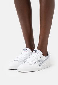 Sneakers bianche con motivo a zigzag nero, suola in gomma, design con lacci e logo sulla linguetta. Materiale in pelle liscia con dettagli perforati.