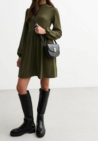 Donna che indossa un abito lungo a maniche lunghe verde oliva, stivali neri fino al ginocchio e una borsa a tracolla nera con dettagli dorati.