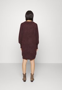 Robe en maille bordeaux à manches longues, coupe décontractée et encolure arrondie, tombant juste au-dessus des genoux. Portée avec des bottines grises.