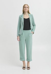 Zelena mint barvana po meri blazer in ujemajoče široke hlače, ki imata teksturirano tkanino, gumbe in zavihke. Črna majica in natikači s trakovi.