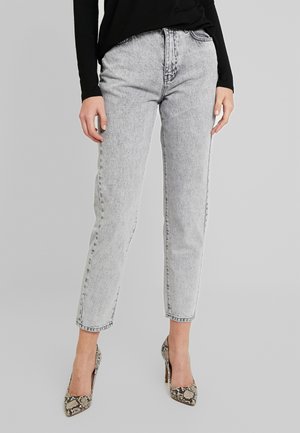 Person trägt hellgraue Acid-Wash-Jeans kombiniert mit einem schwarzen Langarmtop und hochhackigen Schuhen mit Schlangenledermuster und steht vor grauem Hintergrund.