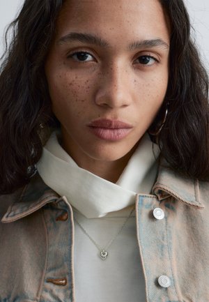 Giovane donna con capelli ricci scuri, lentiggini, che indossa una giacca di jeans chiara, una maglietta bianca, una collana d'argento con ciondolo a forma di cuore e un orecchino a cerchio, rivolta verso l'osservatore.