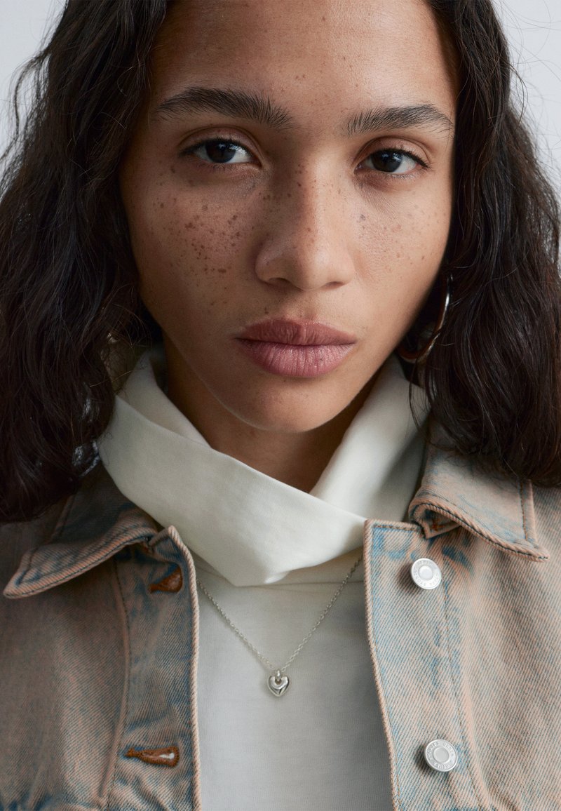 Jeune femme aux cheveux bouclés foncés, avec des taches de rousseur, portant une veste en jean pâle, un haut blanc, un collier argenté en forme de cœur et une boucle d'oreille ronde, regardant devant elle.