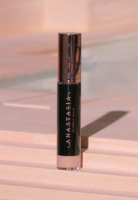 Schwarze Mascara-Tube mit roségoldenem Deckel, versehen mit einem Logo und Text. Der transparente Boden zeigt den Füllstand des Produkts. Glatte, glänzende Oberfläche.