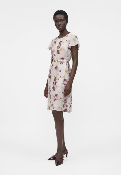 Γυναίκα που στέκεται, φορώντας ένα φλοράλ φόρεμα μέχρι το γόνατο με κοντά μανίκια και λεπτή ζώνη, συνδυασμένο με σκούρα μυτερά τακούνια σε λευκό φόντο.