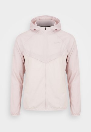 Jasnoróżowa kurtka z kapturem na pełny zamek błyskawiczny, z długimi rękawami i subtelnym logo Nike na piersi, prezentowana na jednolitym białym tle.