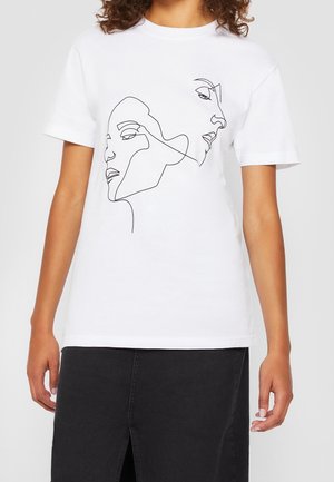 Vrouw die een wit t-shirt draagt met zwarte doorlopende lijntekening van twee overlappende vrouwelijke gezichten, gecombineerd met een donkere rok.