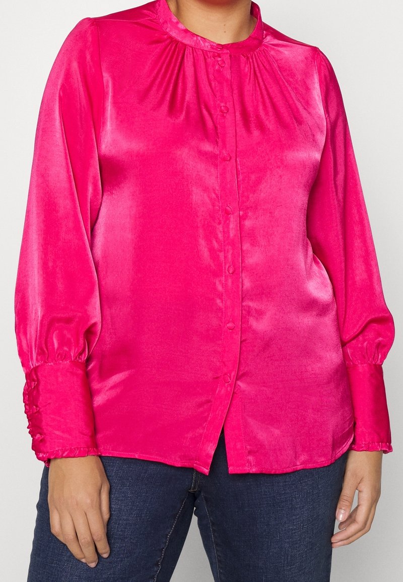 Blouse satinée rose vif à manches longues avec poignets froncés et petits boutons, portée avec un jean en denim foncé.