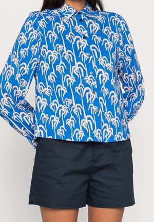 Persoon draagt een blauw overhemd met lange mouwen en witte palmboomprint, gecombineerd met donkerblauwe korte broek.