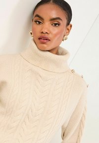 Béžový sveter s pleteným vzorom a vysokým ribovaným golierom, ktorý má detailne gombíky na ramene. Mäkká textúra s hrubým vzorom.