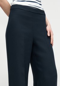 Στενές παντελόνια σε ναυτικό μπλε με απαλό ύφασμα, φαρδύ σχεδιασμό ποδιού και διακριτικές πιέτες. Η ζώνη είναι επίπεδη, χωρίς ορατά μεταλλικά στοιχεία ή σχέδια.